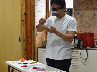 拡大鏡が付いた眼鏡をかけた男性が、長机の前に立ち、ビーズにストラップ部品の取り付けをしている写真