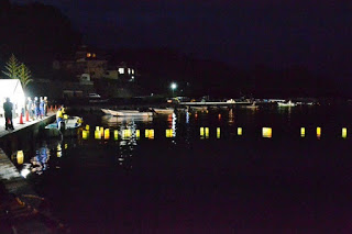 夜の水面に明かりをともした灯ろうが静かに浮かび、岸辺にはそれを見守る人々が集まっている写真