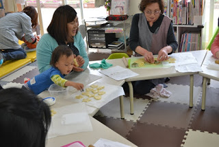 子どもと大人が机を囲んで座り、クッキーの型抜きの作業中で、小さな男の子が型抜きを持ってクッキー生地に手を伸ばしている写真
