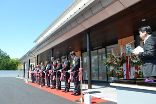 新しく完成した建物の前でスーツ姿の関係者たちが赤いリボンを持って一列に並び、テープカットの式典が行われている写真
