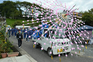 ピンクと白の紙飾りを放射状に広げたような装飾が載った軽トラックが先頭にあり、青いはっぴ姿の人々が行列を作って歩いている様子の写真