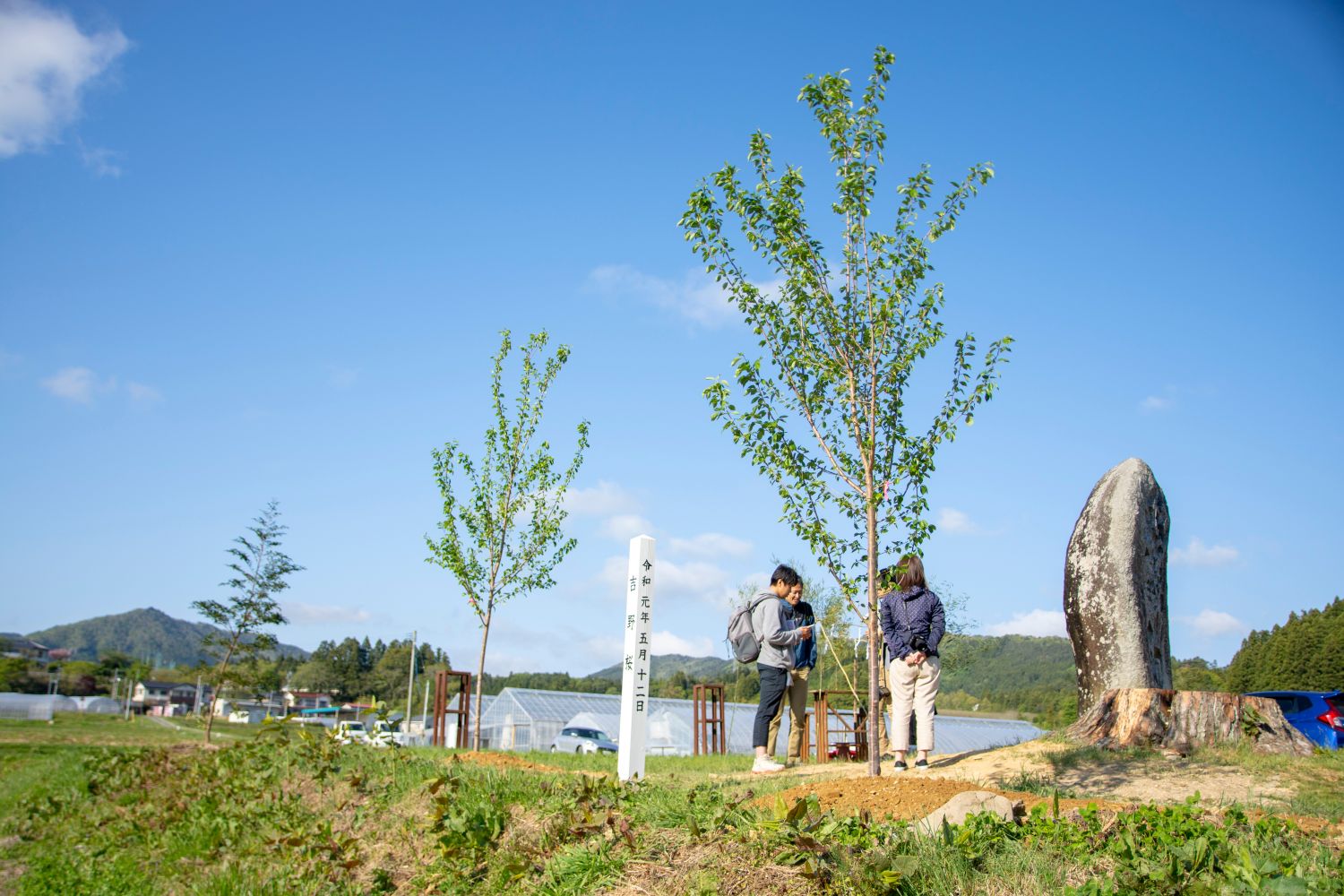 背景には木々やビニールハウス、そして青空の下に広がる山々が見える屋外で、白い縦看板や植樹した木と大きな石碑のそばに4人の人物が立っている写真