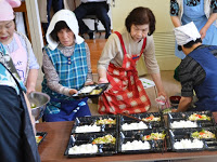 エプロン姿の女性たちが出来上がったお弁当のおかずやごはんを容器に盛り付けて配膳している写真