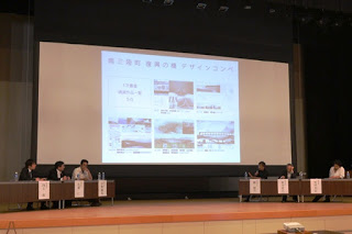 ステージ上に6人のパネリストが横一列に座っており、背後のスクリーンには「南三陸町復興の橋デザインコンペ」と書かれたスライドが映し出されている会議の様子が写っている写真