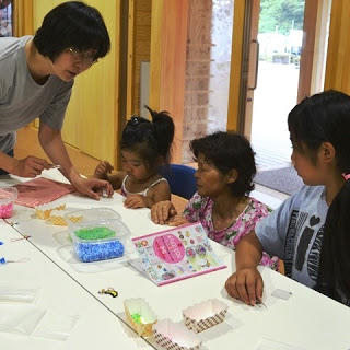 女の子と高齢の女性がビーズやクロスステッチの本が置かれたテーブルに向かって座り、鈴木さんが指導をしている様子の写真