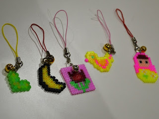 テーブルの上に5つのアイロンビーズで作られたストラップが並んでおり、左から緑色のハート、黄色と黒の三日月、ピンクの中にチューリップ、黄色とピンクのひよこ、ピンクと黄緑色の人形にそれぞれ異なる色の紐が付いている写真