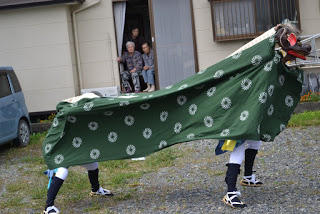 緑色の布をかぶった獅子舞の形をした大きな衣装を二人が動かしている様子の写真