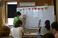 講師がホワイトボードに吊るした布細工を使って説明し、自席に座った参加者も同じようなものを手に取りながら作業している写真