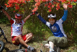 赤いツツジの下にマウンテンバイクを置き、ヘルメットを被った二人の選手が両手を挙げて笑顔で達成感を味わっているゴール直後の写真