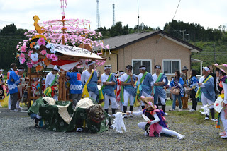 屋外で花の飾りが施された神輿の前に青い衣装の人々が集まり、子どもが踊っている様子の写真