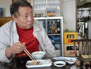 食堂で赤いシャツに上着を羽織った男性が、貝やウニが置かれたテーブルで白いごはんとおかずを食べている様子の写真