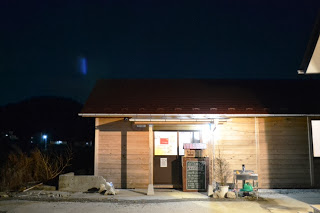夜の暗がりの中、明かりの灯った木造の建物の入り口が見え、入口横に手書きの黒板看板が置かれている自習支援TERACOの外観写真