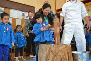 青いスモックを着た園児が、木の杵(きね)を使い大人の女性がしっかりと手を添えてサポートしながらもちつきをしている写真