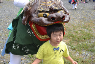 獅子舞の頭の衣装をかぶった人のすぐそばで笑顔の幼児が立っている様子の写真