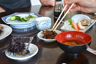 テーブルに小皿に盛り付けられた1ケの殻付きウニ、みそ汁、お刺身、きゅうりの漬物などが並べられ、割りばしで小鉢にはしをつけている写真