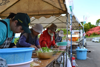 複数の女性がテントの下で、それぞれまな板やボウルを使いながら野菜を切ったり混ぜたりしていてしたごしらえをしている、大会の食事提供準備の写真