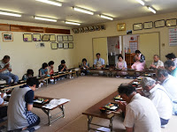 ロの字型に長机が設置された畳の部屋で、手作りのお弁当を食べながら和やかに過ごしている参加者の写真