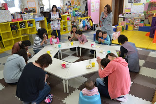 六角形に並べられたテーブルの周りに大人と子どもたちが座り、クッキーが乗った皿と飲み物のコップを前に手を合わせ、いただきますのポーズをしている写真