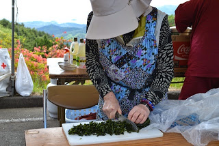 日よけ帽子とエプロンを着けた女性が、屋外のテントの下でまな板の上の野菜を包丁で細かく刻んでいる、大会の調理準備の写真