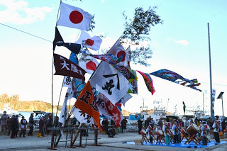 漁港の岸壁に多数の大漁旗や日本国旗がはためき、太鼓演奏をする人々や観客が集まっている寄木ささよの様子の写真