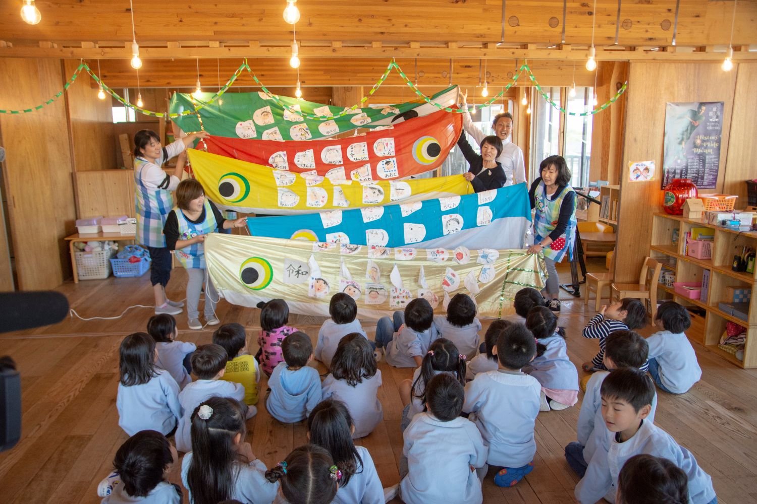 室内で先生たちが5色の鯉のぼりを披露し、床に座った子どもたちが注目している写真