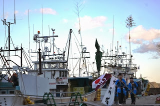 大漁旗を持った法被姿の男の子たちが、停泊している漁船の横を歩いている様子の写真