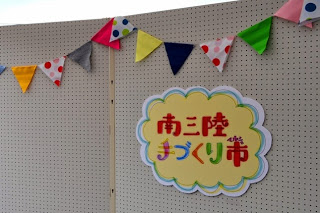 南三陸手づくり市と書かれた黄色い雲形のプレートが、色とりどりの三角の旗飾りと一緒にボードに飾られている写真