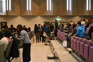 紫色の座席が並ぶ室内のホールで、多くの人が座ったり立ち歩いたりしながら開演前の準備や会話をしている様子が見られる写真