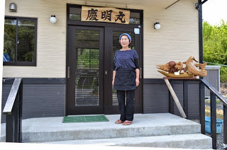 女性が和風の建物の玄関前に立っていて、頭に青い手ぬぐいを巻き、黒いエプロンを身につけ、正面を向いて穏やかに微笑んでいる写真