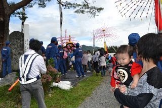 青い法被を着た男性や観光客が狭い通路を進み、手前には赤い衣装を着た幼児を抱く女性が立っている秋祭りの行列の写真