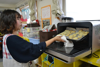 オーブンの扉を開けて、布を使ってクッキーの乗ったトレイを持ち、焼き上がったクッキーを取り出している女性の写真
