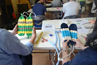 左側の席の参加者は黄色や緑色に灰色、右側の席の参加者は紺色と水色に白色の底面の土台部分が完成し、両手で持って見つめている写真