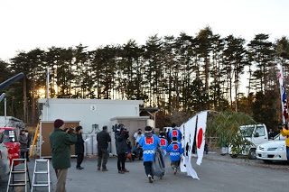 カメラやビデオを撮影している観衆が見守る中、大漁旗を持った法被姿の男の子たちが走り出している後ろ姿の写真