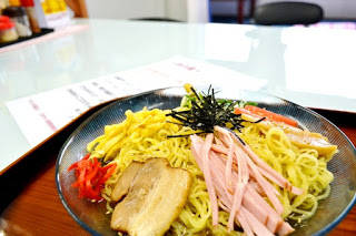 白いガラスのテーブルに置かれた冷やし中華の皿には、錦糸卵、ハム、チャーシュー、紅しょうが、きゅうりなどが盛りつけられている写真