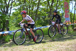 木漏れ日の中、「たつがねMTB」と書かれたのぼりが立つコースを、2人の子どもたちが先頭を競いながら走っている写真