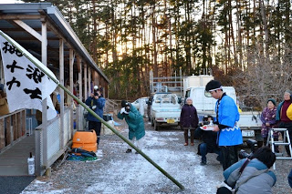 仮設住宅の前で大漁旗を掲げ、法被を着た畠山史也君がお神酒を注いている寄木ささよの様子の写真