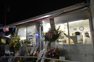 ガラス張りのお店の外観が夜に明るく照らされ、店内にはスタッフの姿が見え、店頭には開店祝いの花がずらりと並んでいる写真