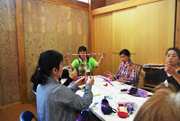 講師が両手でカラフルな色の長い布ひもを持ち上げて見せながら、4人の参加者と一緒に机を囲んで布ぞうり作りを行っている写真