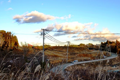 起伏のある造成地に沿って道路が蛇行し、遠くに工事車両や空が広がる夕暮れの造成地の写真