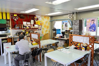 壁にポスターや装飾が飾られた明るい照明の下、白いテーブルと椅子が並び、2人のお客さんが座っている飲食店内の写真