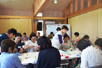 十数人の参加者が大きな教室の中で布ぞうりを作っており、講師やスタッフの方が机の間を回って指導している写真