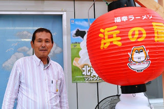 「福幸ラーメン浜の屋」と書かれた赤い提灯の横で、笑顔の男性が立っている写真