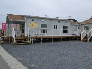 白いウッドデッキが特徴の一軒家風プレハブに「プラス」と書かれた看板が掲げられている美容室の外観の写真