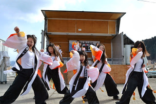 ステージ前で白とオレンジの法被を着た女性たちが力強く踊る姿をとらえた、演舞パフォーマンスの写真