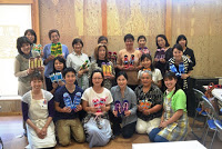 完成した布ぞうりを手に持った参加者たちが笑顔でカメラに向かって並んで座り、教室内で記念撮影をしている写真