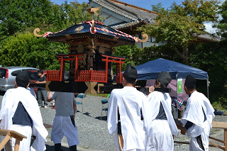 黒い烏帽子と白装束の男性数人が赤黒く装飾された神輿を担ぎ、青いテントが立つ神社境内を進んでいる秋祭りの神輿渡御の写真