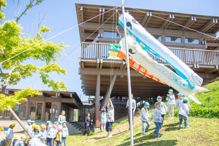 段差のある土地の上下に建つ建物前の芝生の斜面に立ったポールにカラフルなこいのぼりを掲げている様子を下に集まったたくさんの園児たちが眺めている様子の写真