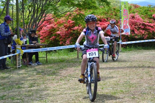 背景に赤いツツジと「たつがねMTB」と書かれたのぼりが立つ山道で、ゼッケン191番をつけた笑顔の沼田素直さんがゴールに向かってマウンテンバイクをこいでいるレース中の写真