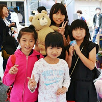 4人の子どもたちが一緒に立っていて、うち3人は手でピースサインをしており、1人は大きなぬいぐるみのクマを抱えている写真