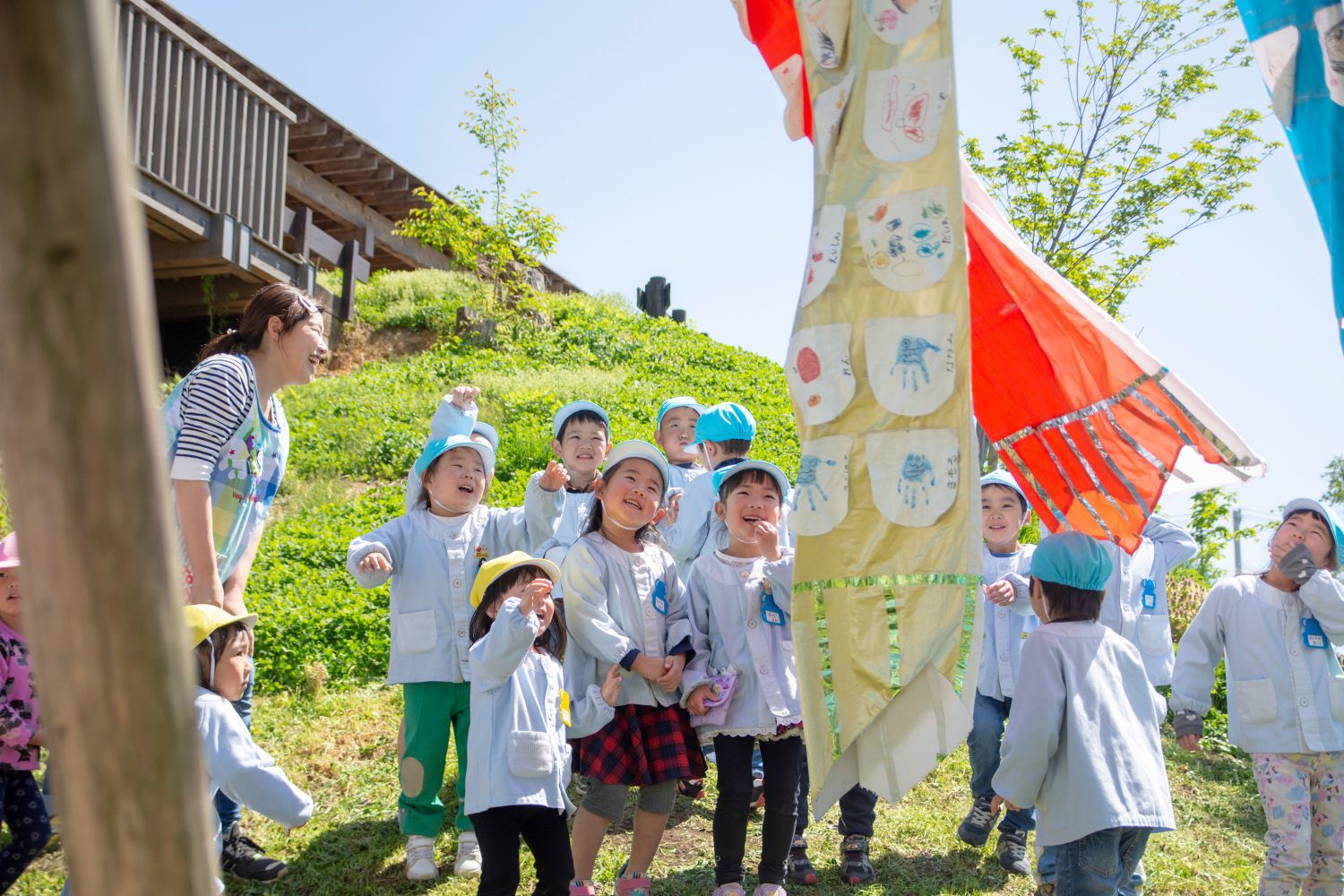 屋外で保育士とともに笑顔の子どもたちが楽しそうに風に揺れる鯉のぼりを見上げている写真